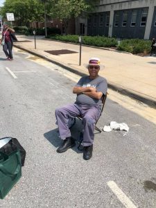 Benny sits on a folding chair on the street, watching volunteers create T2T's chalk drawing