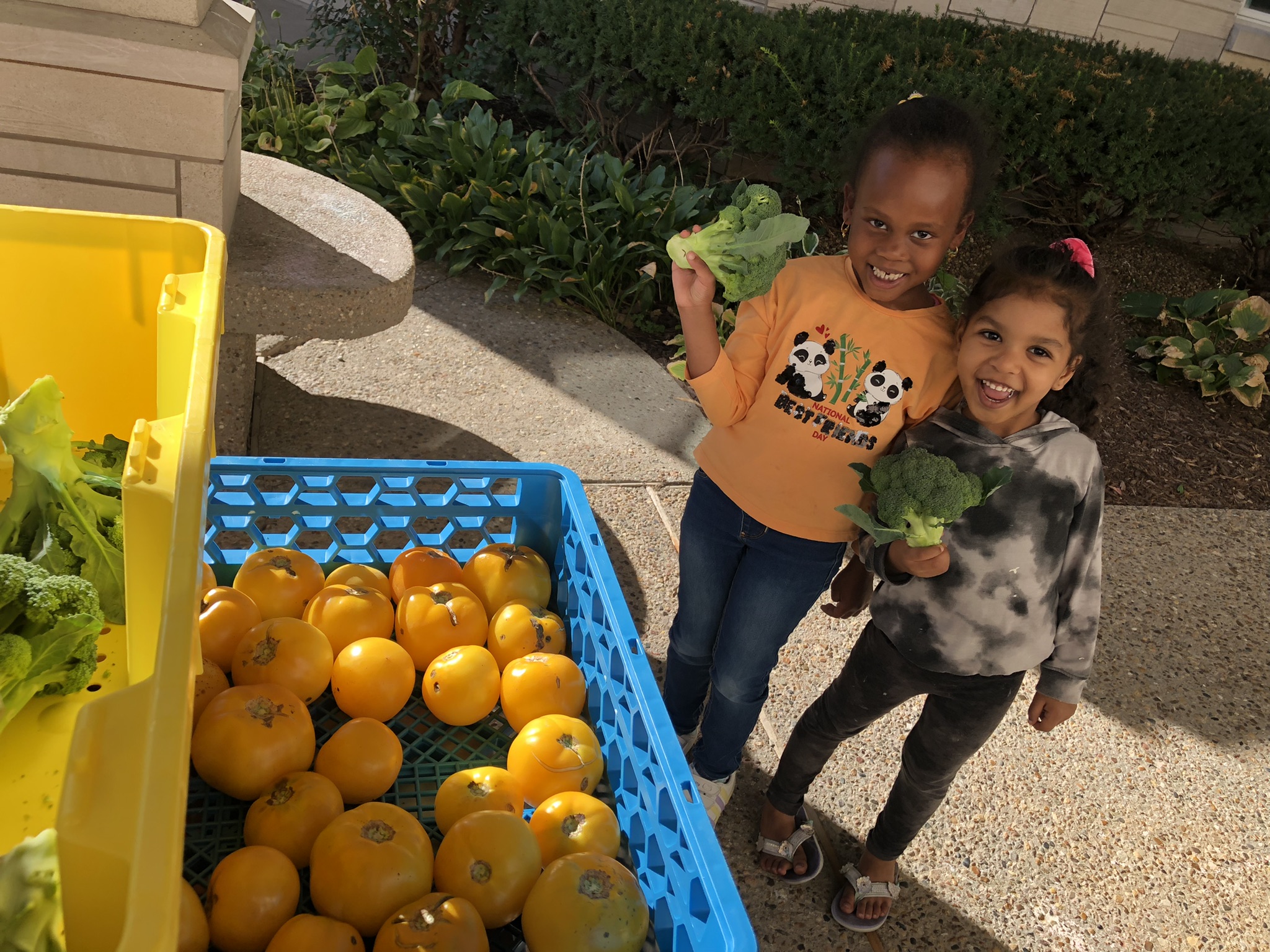 Two young girl smile wide as they hold broccoli at a free produce stand