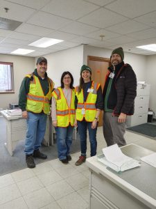 Four people, most wearing bright yellow vests, stand in an office space