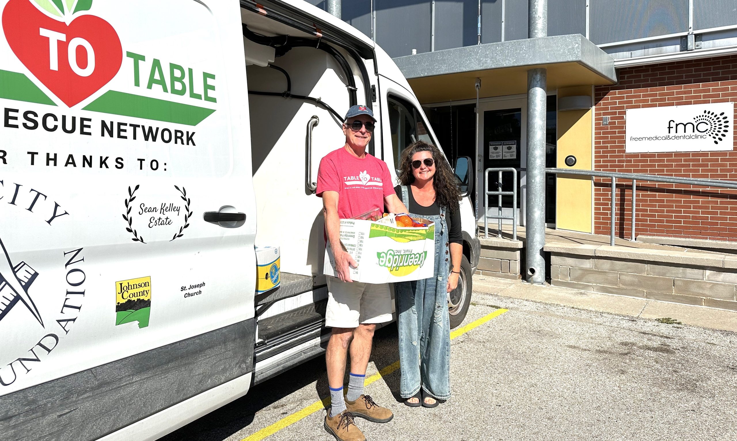 A T2T volunteer, holding a box of vegetables, and a staff member with Free Medical Clinic pose with the T2T van in front of the Free Medical Clinic building.