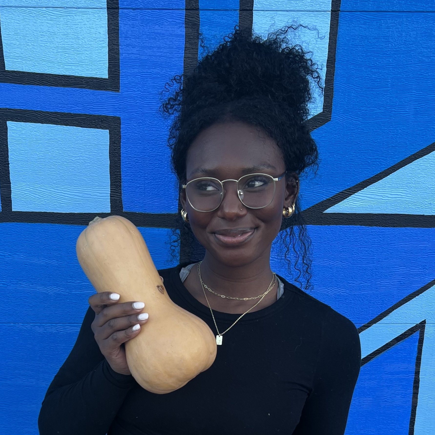 Jada stands against a blue wall and holds a winter squash in her hand.
