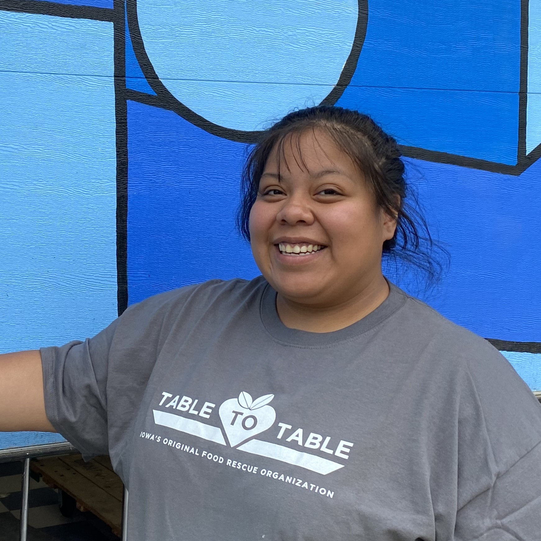 Yesenia, wearing a gray shirt that says Table to Table, stands against a bright blue wall.