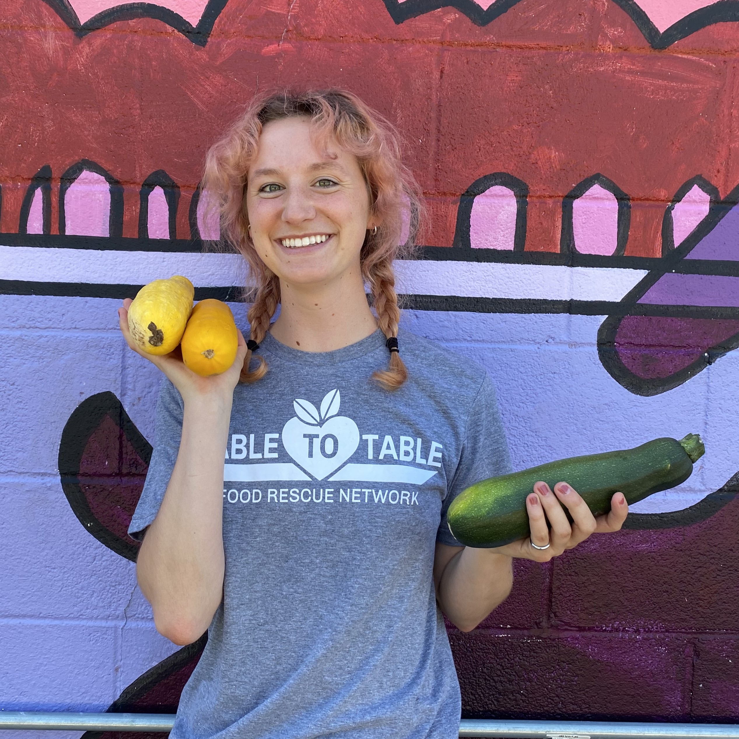 Emma stands against a purple painted mural holding yellow and green zucchini