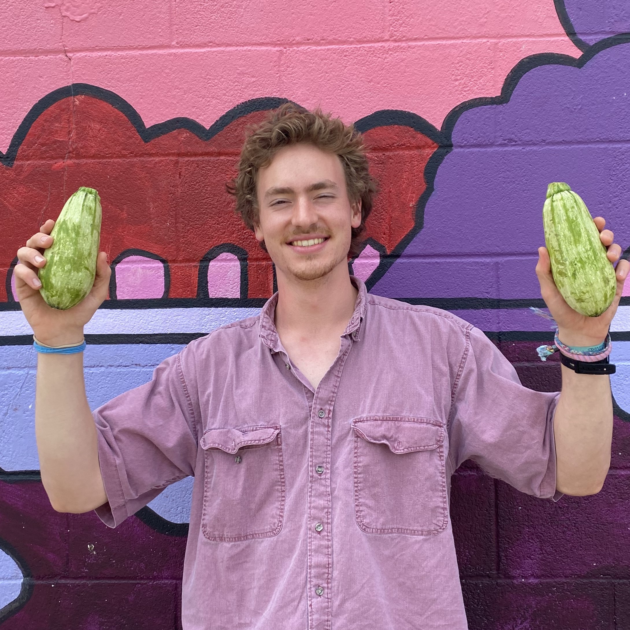 Adam stands against a purple painted mural, holding two green zucchini