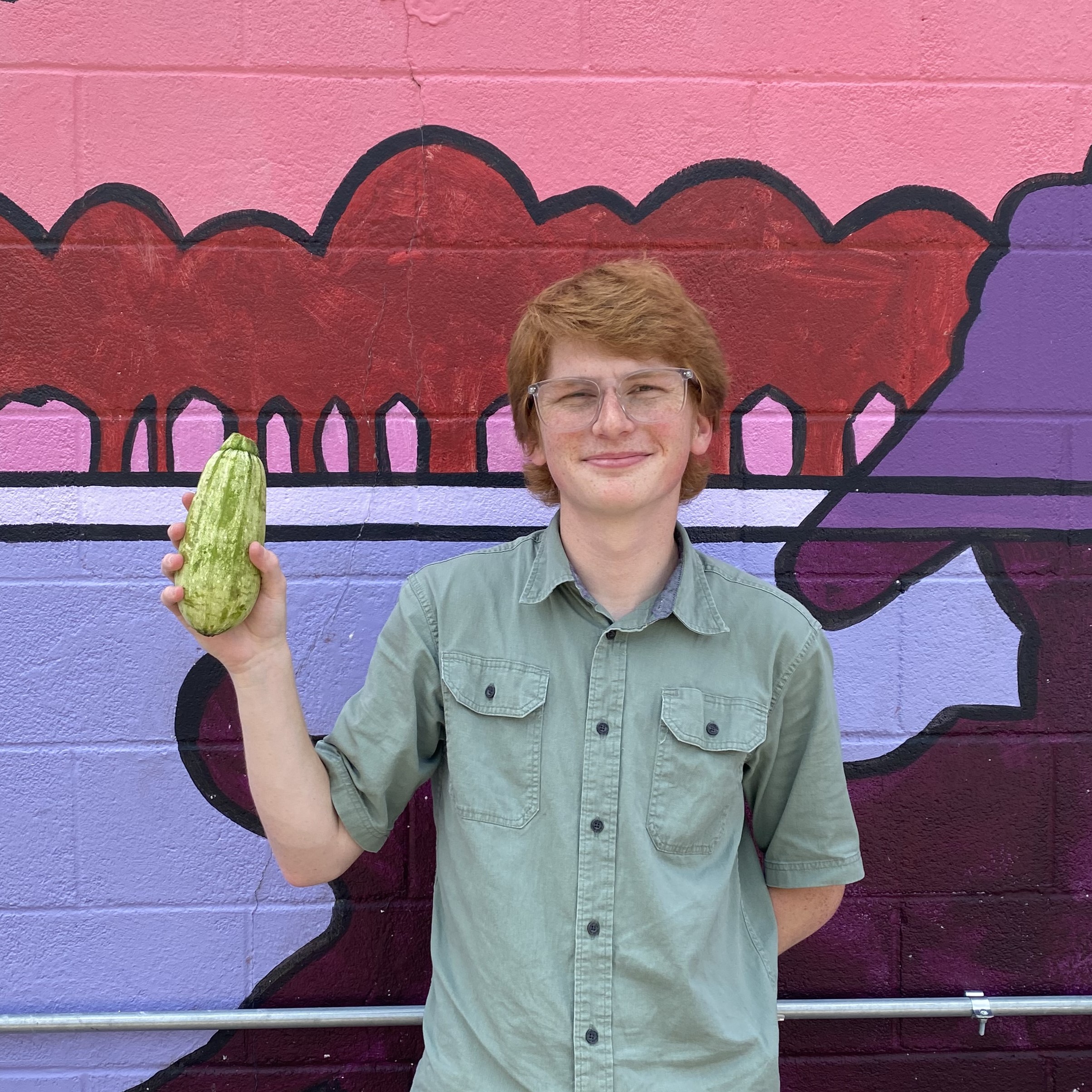 Noah stands against a purple painted mural, holding a green zucchini